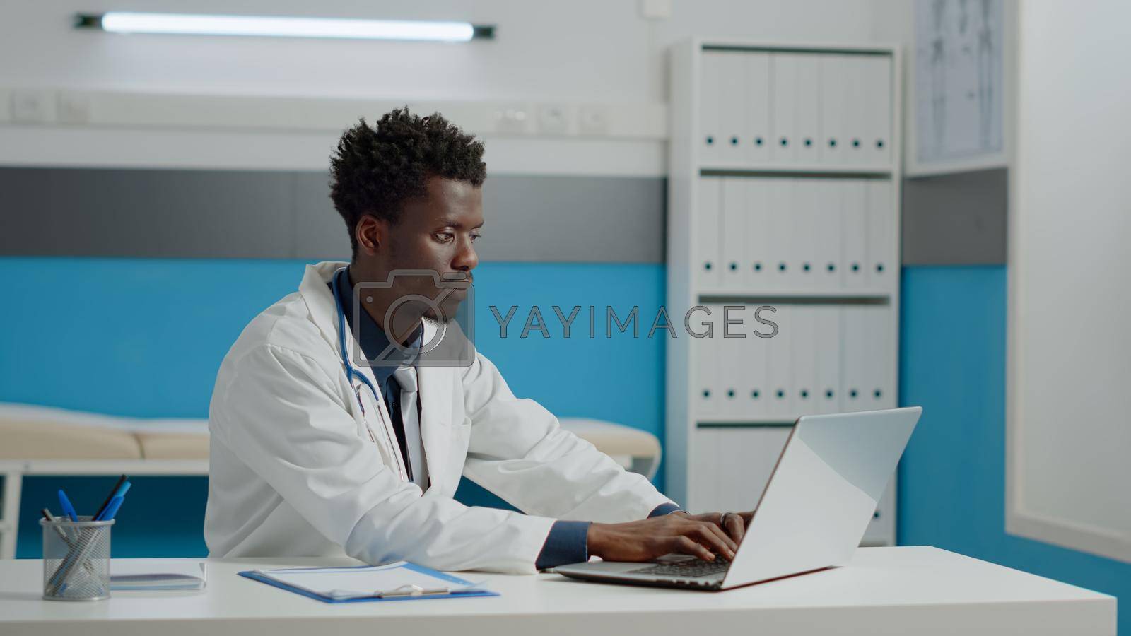 Portrait of healthcare specialist looking at camera while smiling in medical cabinet. Young medic with healthcare uniform typing on laptop keyboard and sitting at desk in doctors office