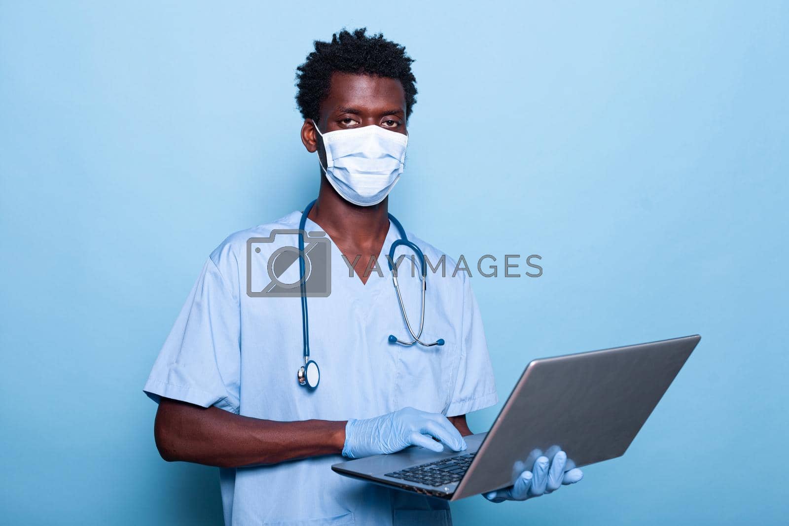 Healthcare specialist with laptop in hand looking at camera. Medical nurse with stethoscope and blue uniform wearing face mask against coronavirus pandemic. Assistant with gloves