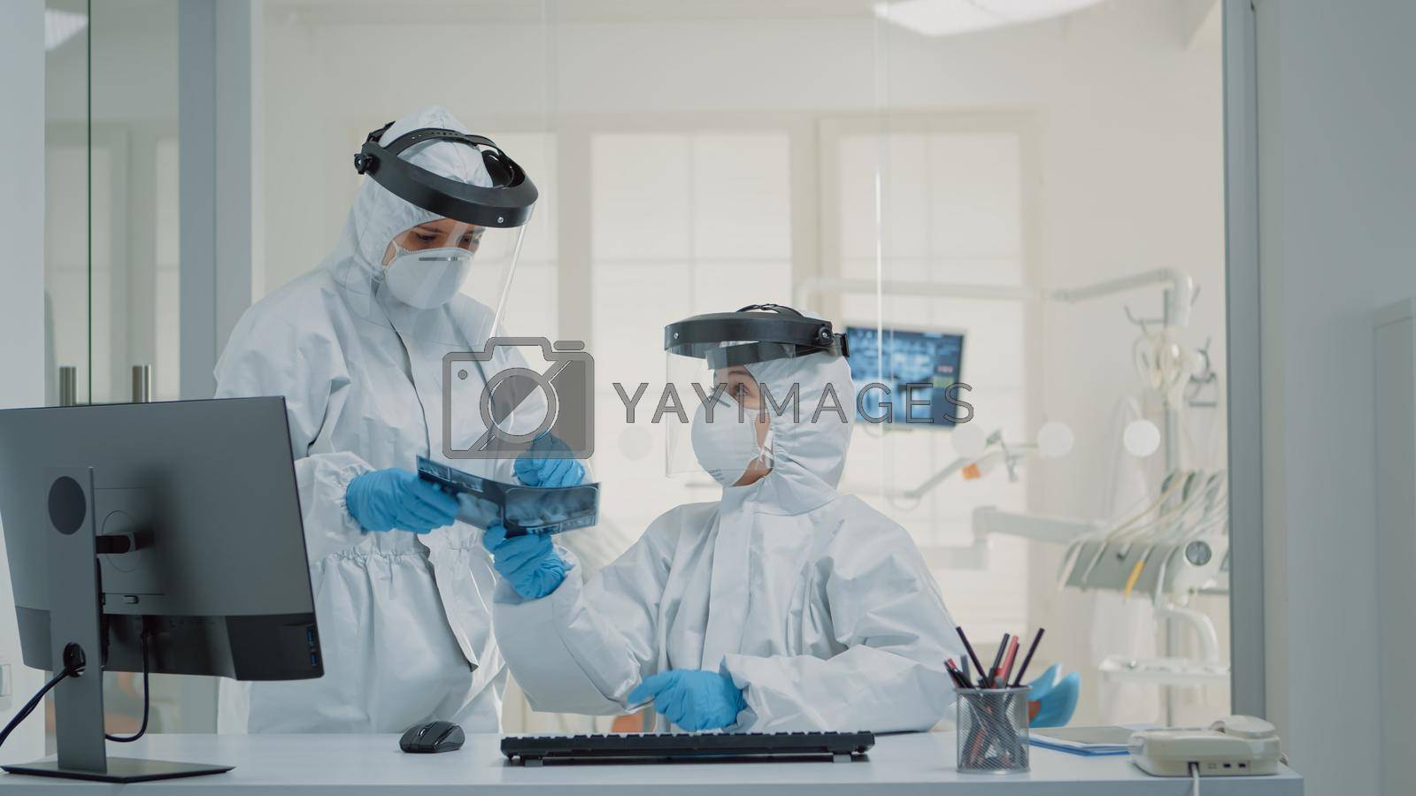 Medical stomatology staff working on teeth healthcare for patient at dental clinic, wearing protection suits. Nurse using modern computer at desk while dentist holding x ray for implant