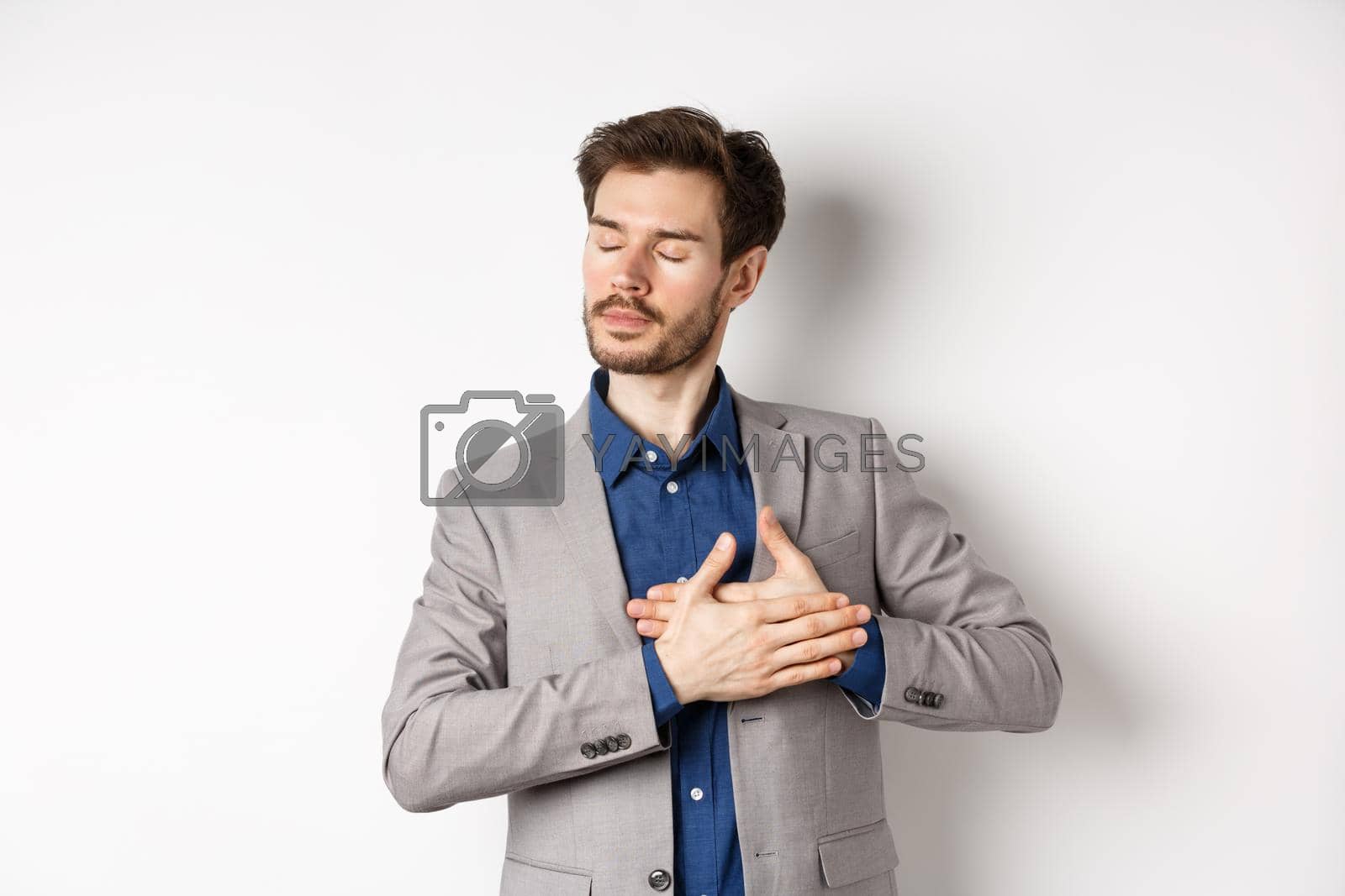 Royalty free image of Romantic bearded man in stylish suit holding hands on heart, close eyes and remember something with tender feelings, standing nostalgic on white background by Benzoix