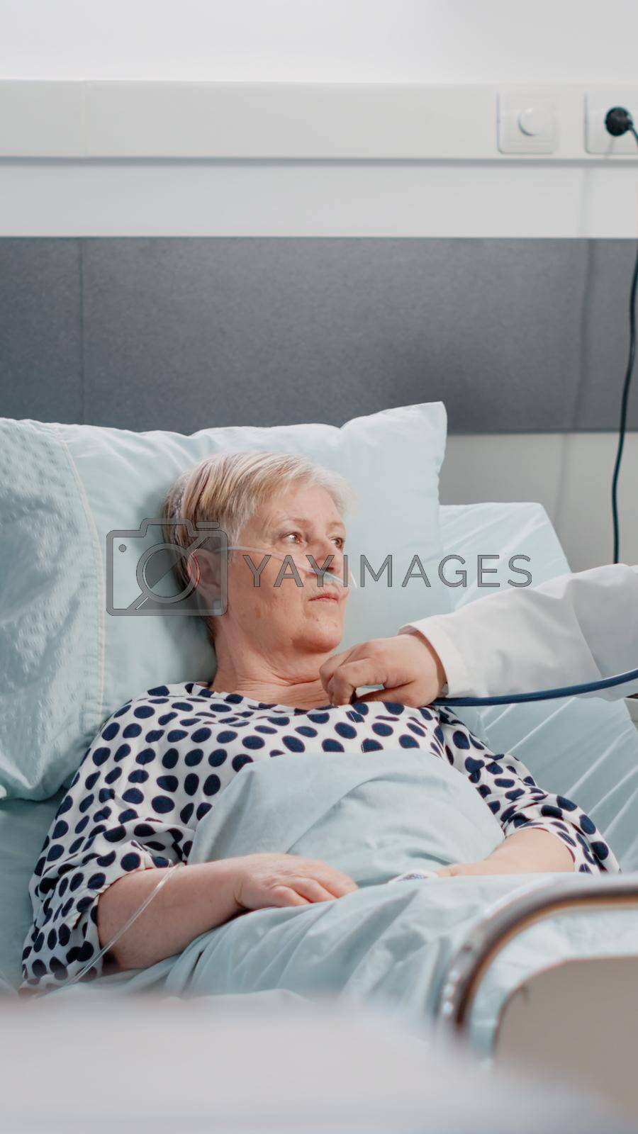 Medical team doing healthcare consultation for senior patient with illness. Doctor using stethoscope to examine sick woman with nasal oxygen tube in bed while nurse giving assistance