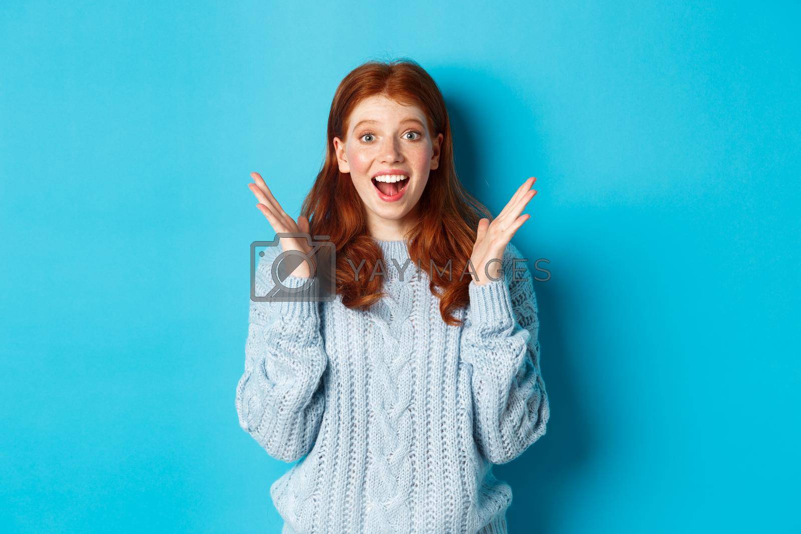 Royalty free image of Surprised and happy redhead girl clap hands and staring at camera, smiling amazed, standing against blue background by Benzoix