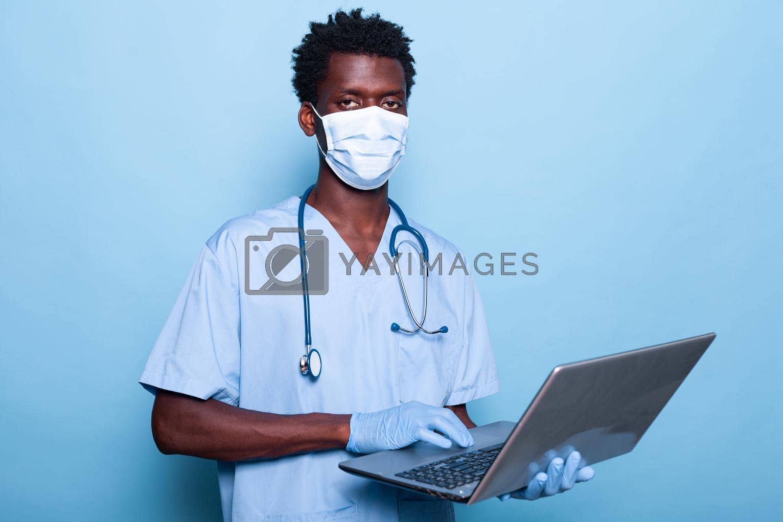 Healthcare assistant with laptop in hand wearing face mask. Medical nurse with uniform and gloves for coronavirus pandemic protection holding device. Specialist with stethoscope