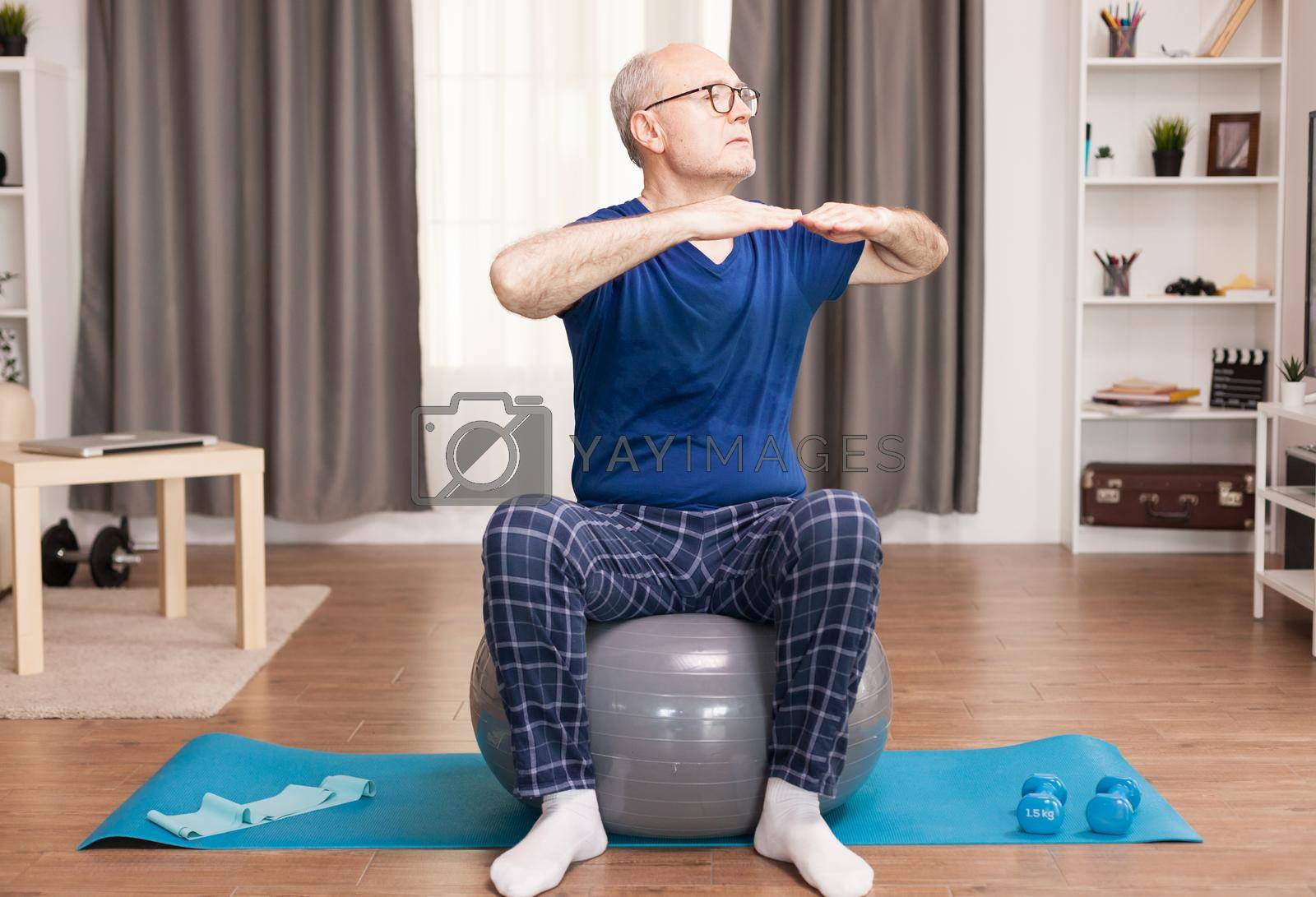 Active old man doing sport in his cozy apartment using swiss ball and yoga mat. Old person pensioner online internet exercise training at home sport activity with dumbbell, resistance band, swiss ball at elderly retirement age.
