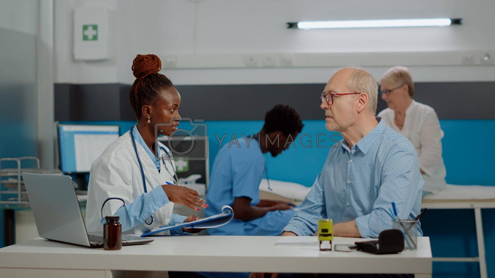 Healthcare specialist discussing with old patient at desk in medical office. Doctor checking disease symptoms for senior person while nurse and elder woman sitting in background