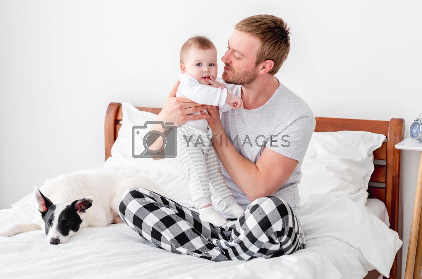 Young father sitting in the bed with little son and cute dog in the room with sunlight and hugging his baby boy. Man staying in bedroom with his child and pet. Guy with kid and doggy in morning time