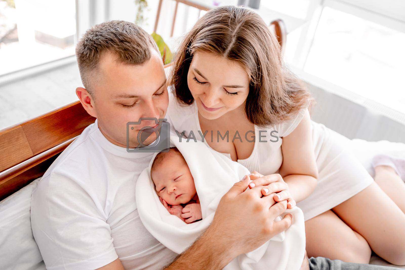 Beautiful young family with newborn daughter in the bed with daylight. Adorable infant baby girl sleeping in hands of her mother and father at home