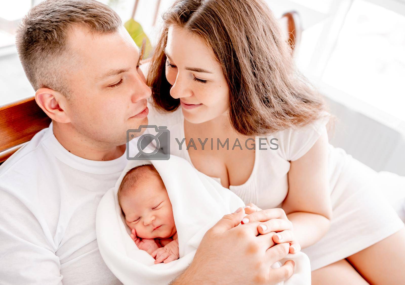 Beautiful young family with newborn daughter in the bed with daylight together. Adorable infant baby girl sleeping in hands of her mother and father at home, and parents looking and smiling