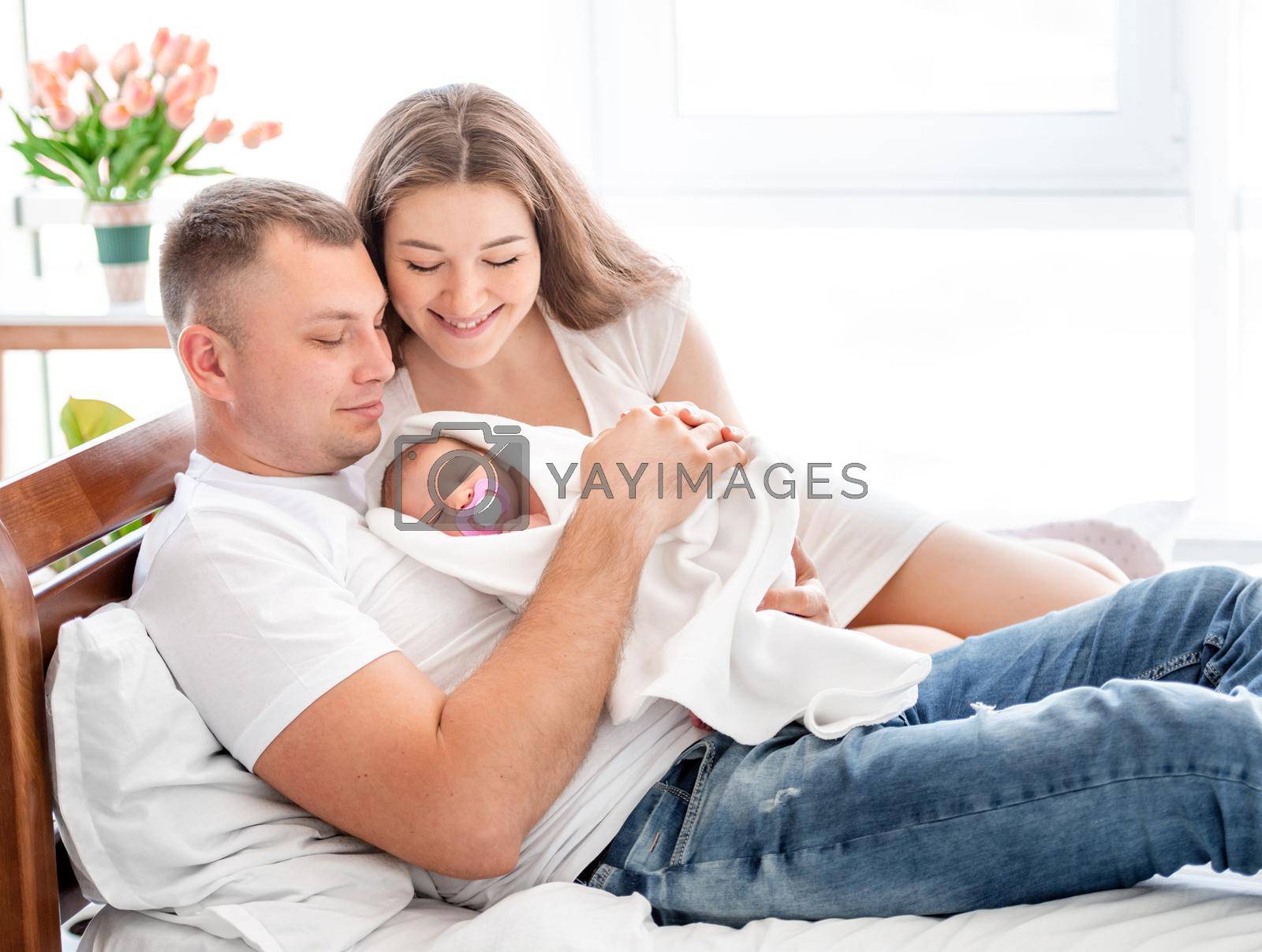 Young parents lying in the bed with newborn daughterand smiling. Adorable infant baby girl sleeping in hands of her mother and father at home