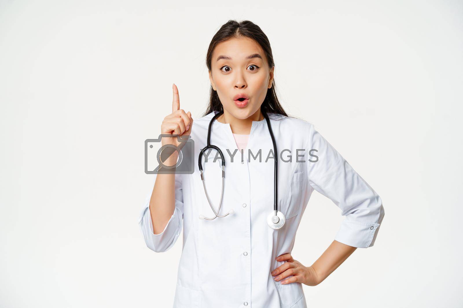 Royalty free image of Healthcare and medical concept. Asian woman doctor raising finger, found solution, treatment for patient, pointing up and looking enthusiastic, white background by Benzoix