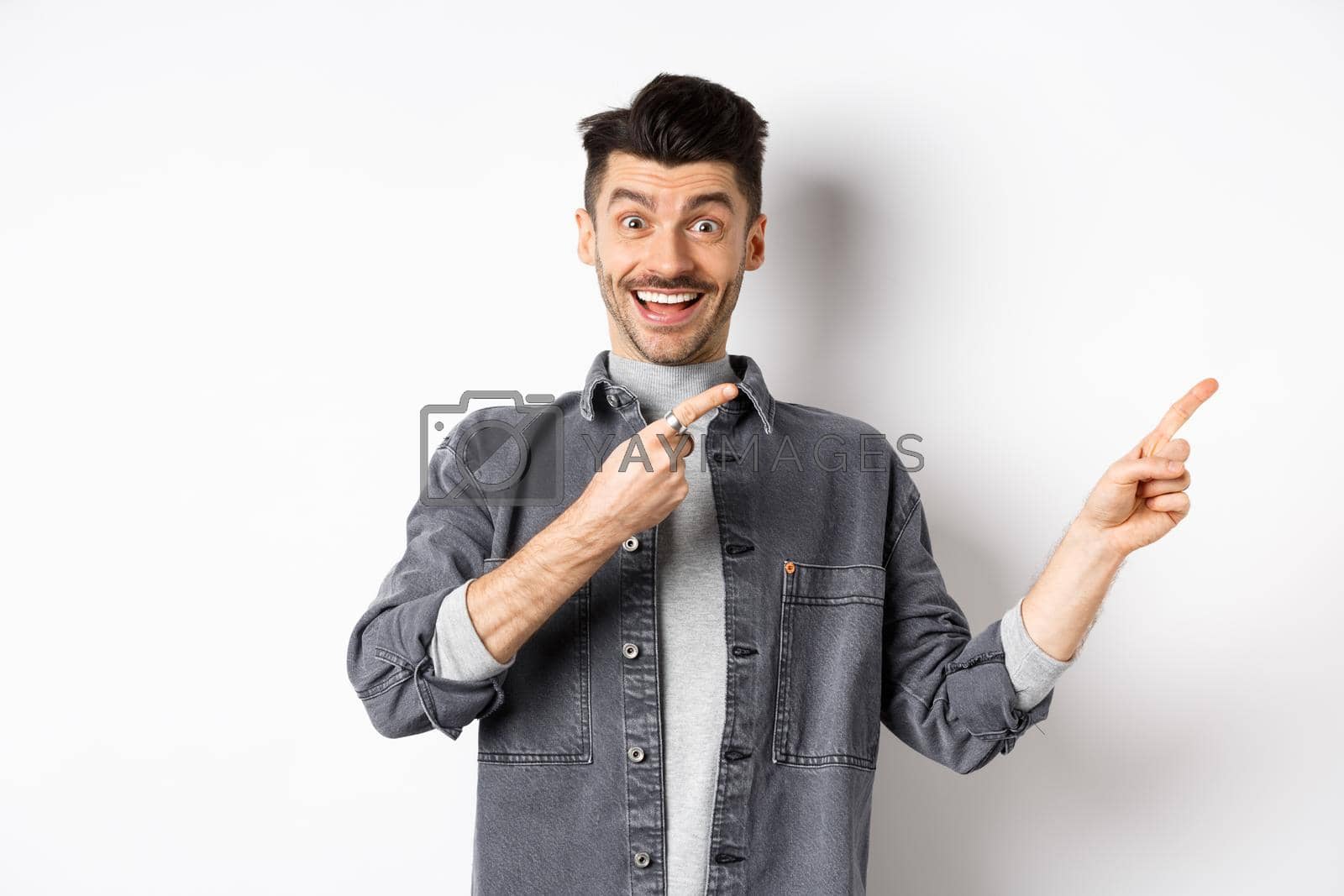 Royalty free image of Look here. Happy stylish man smiling, pointing fingers right at empty space, showing cool promo offer, standing in denim jacket on white background by Benzoix