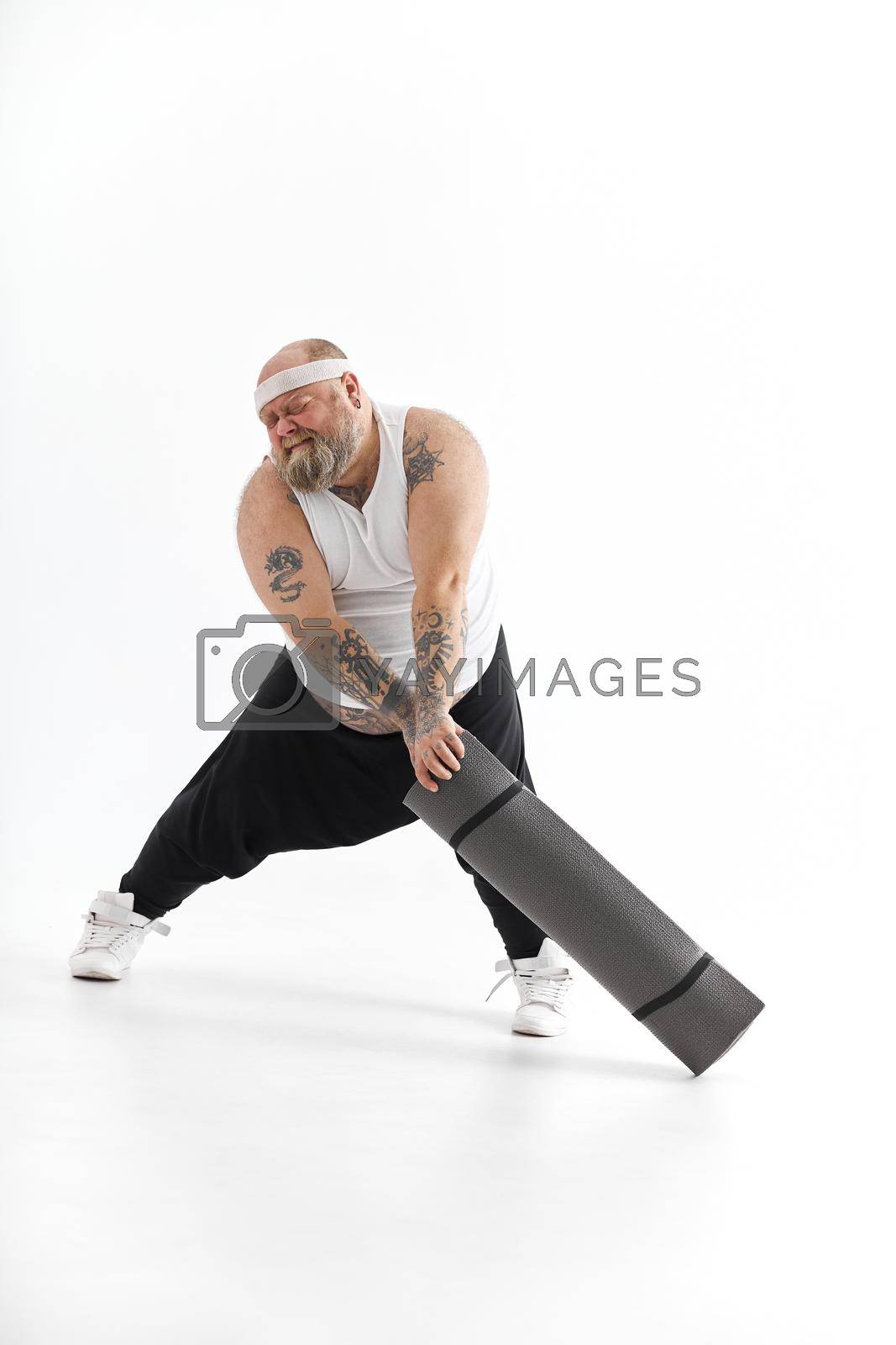 Royalty free image of Happy fat man with big belly and tattos in sports wear with exercise mat by Yaroslav_astakhov