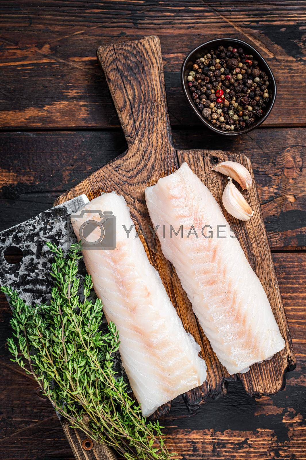 Royalty free image of Raw cod loin fillet steak on wooden board with butcher cleaver. Dark wooden background. Top view by Composter