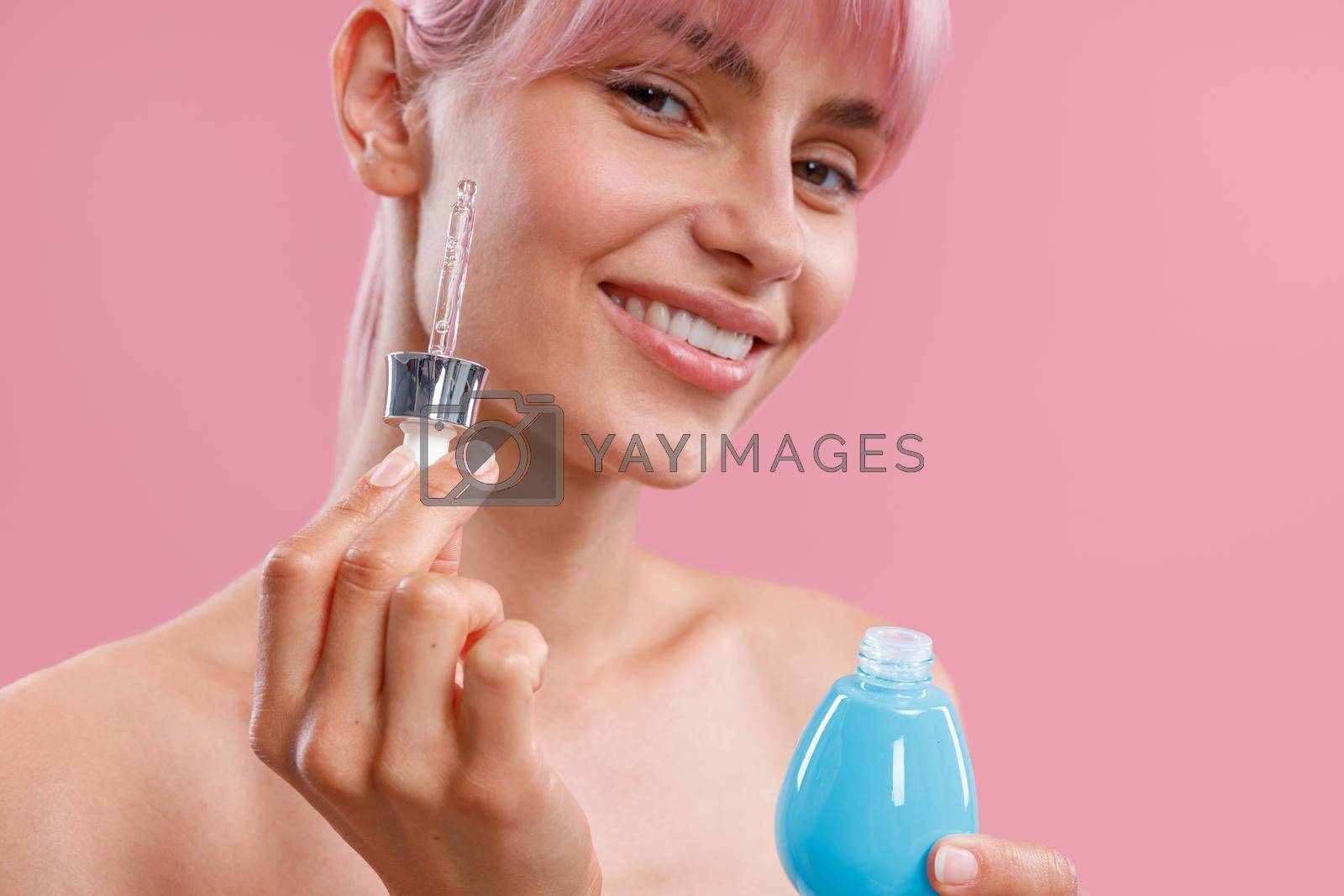Royalty free image of Portrait of young woman with pink hair holding dropper and a bottle of serum or hyaluronic acid, posing isolated over pink background by Yaroslav_astakhov