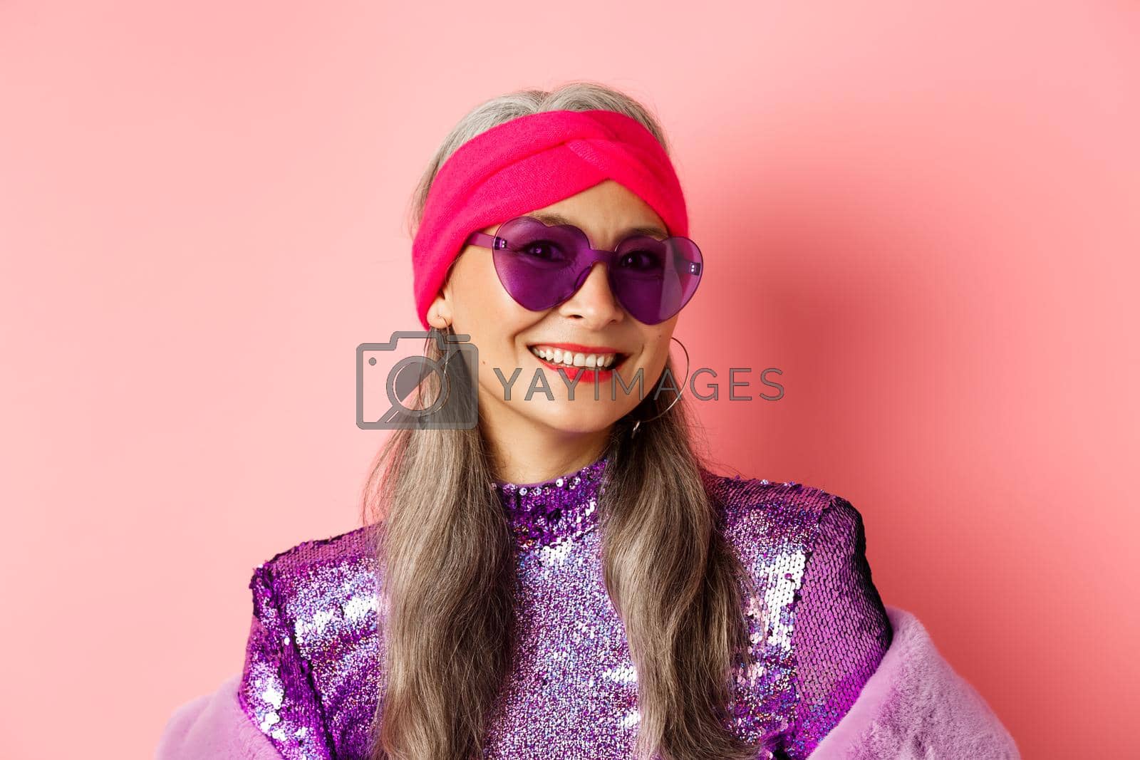 Royalty free image of Fashion. Close up of stylish senior woman smiling happy at camera, wearing sunglasses and disco headband, pink background by Benzoix