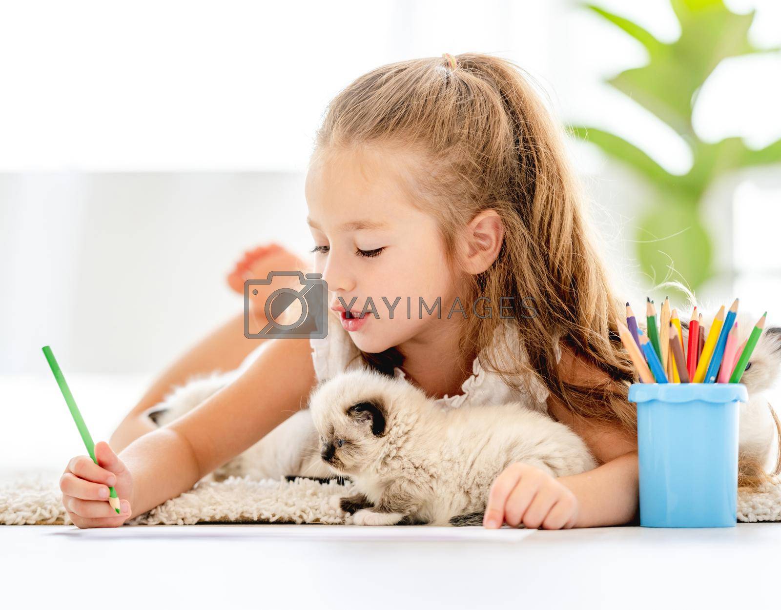 Child girl painting with ragdoll kittens and lying on the floor. Little female person drawing with colorful pencils and kitty pets close to her at home