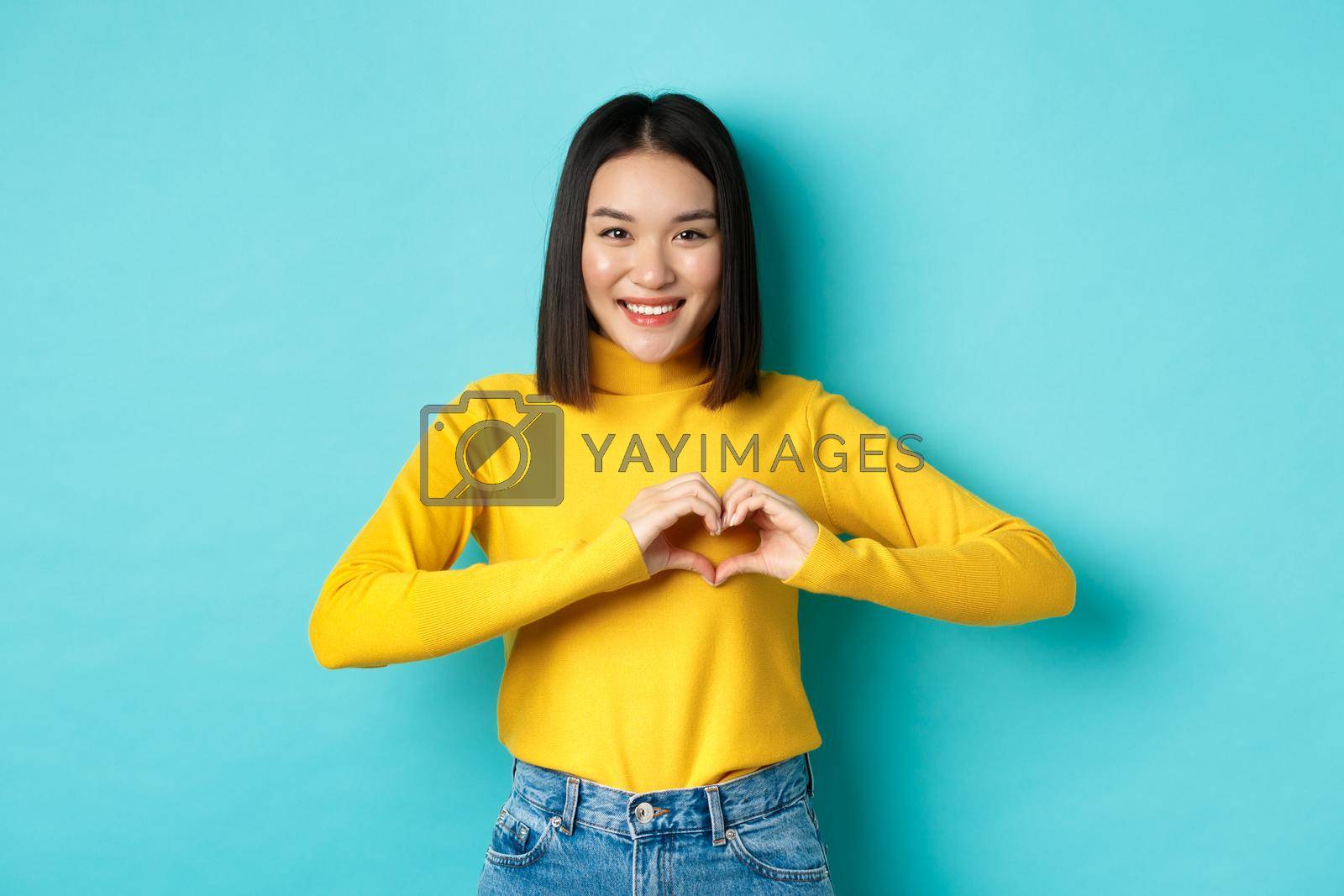 Valentines day and romance concept. Beautiful asian woman show I love you, heart gesture and smiling, standing against blue background.
