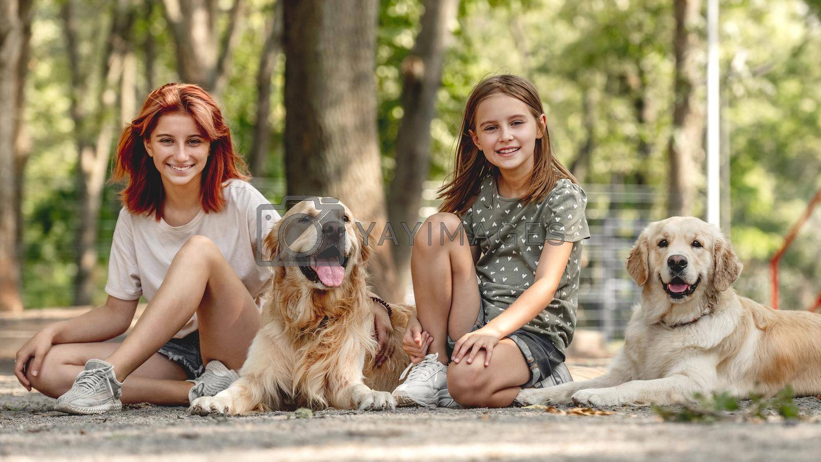 Girls with golden retriever dogs sitting in the park. Sisters with doggy pets outdoors looking at camera