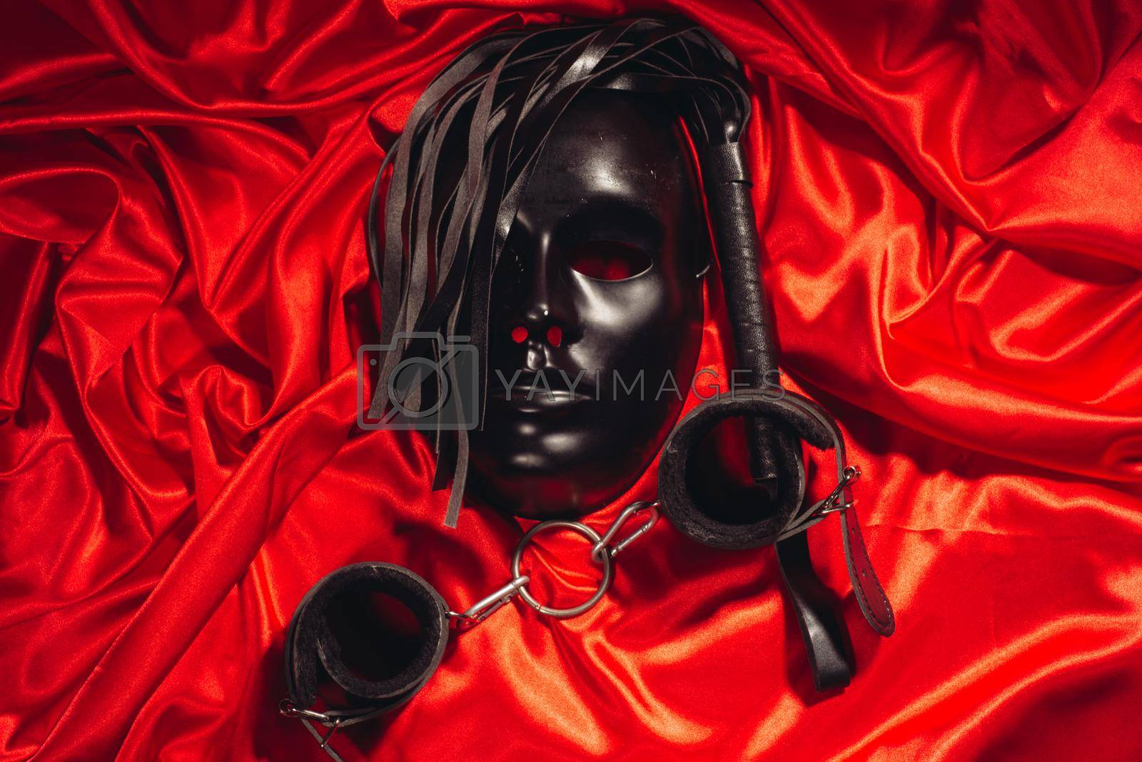 Bondage, kinky adult sex games, kink and BDSM lifestyle concept with a mask, pair of leather handcuffs, flogger, ball gag and a coller with a leash attached on red silk with copy space