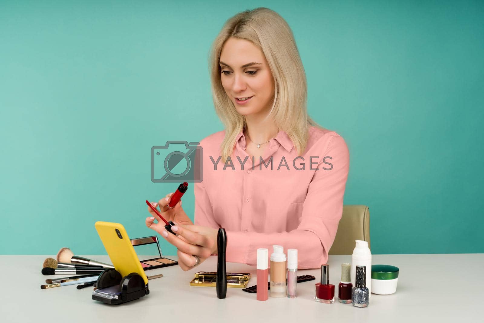 Candid shot of cute young Caucasian woman blogger presenting beauty products and broadcasting live video to social network, using lip gloss while recording everyday make up tutorial