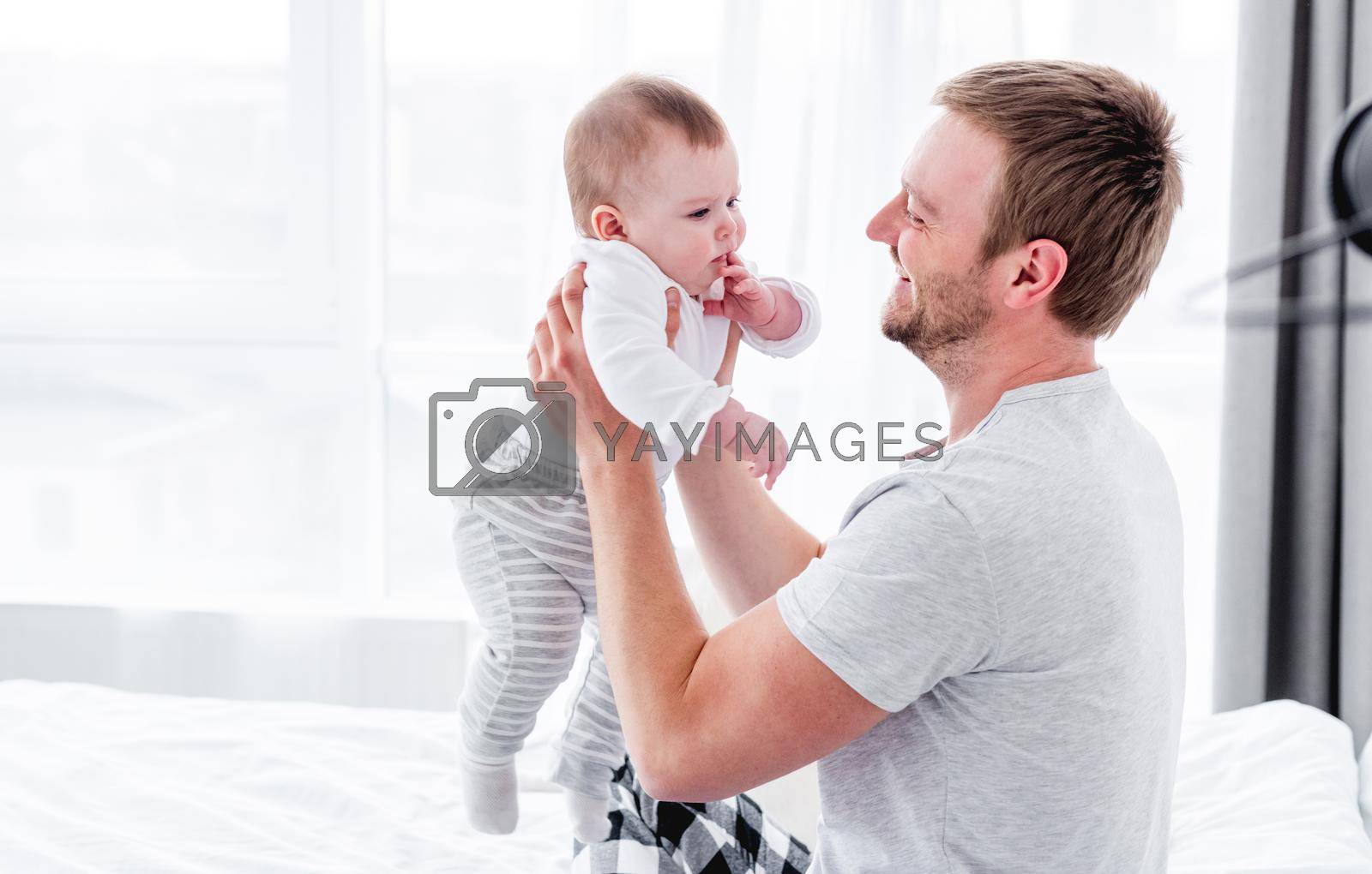 Closeup portrait of young father sitting in the bed and holding little son in his hands in the room with sunlight. Man staying in bedroom with his child. Guy with kid in morning time