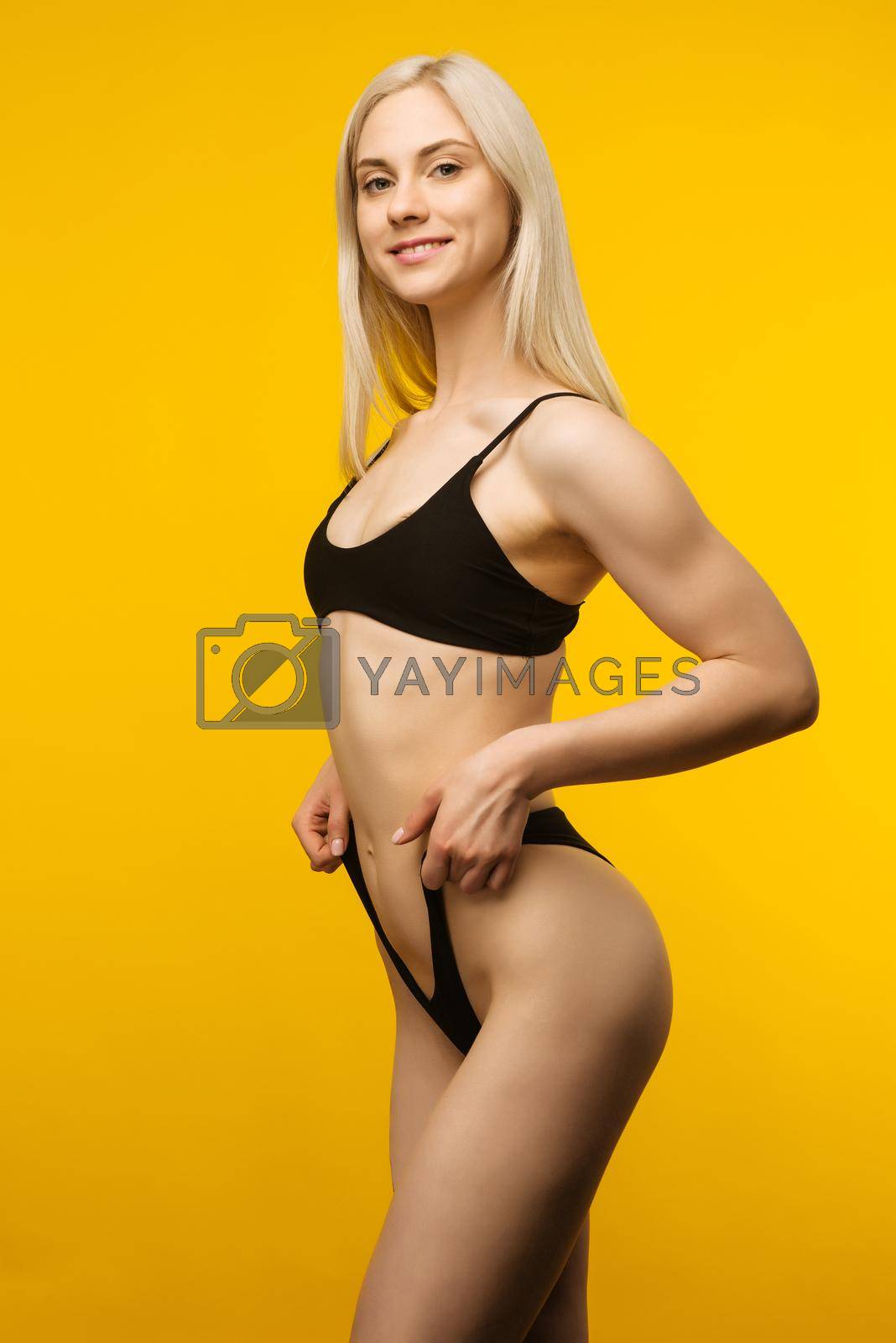 Laughing sporty girl in black bikini posing on yellow background. Photo of attractive girl with slim toned body. Beauty and body care concept