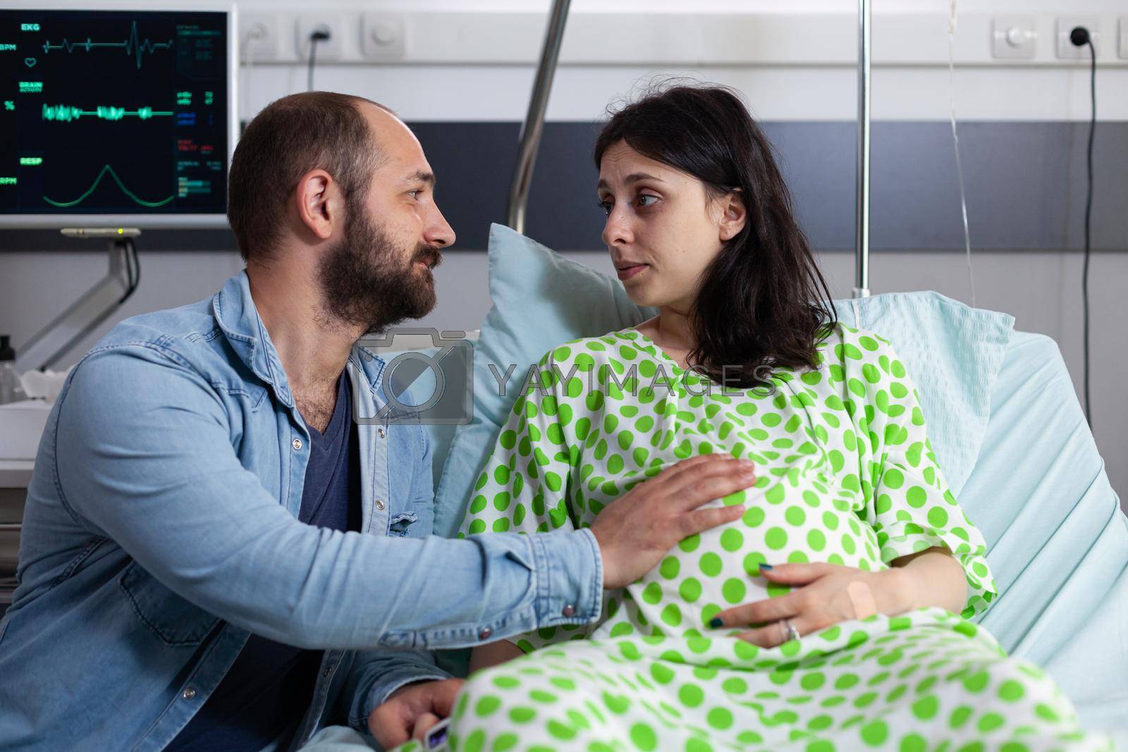 Couple expecting baby at healthcare facility in hospital ward. Father of child holding hand on belly of pregnant woman sitting in bed. Young parents preparing for childbirth and parenthood