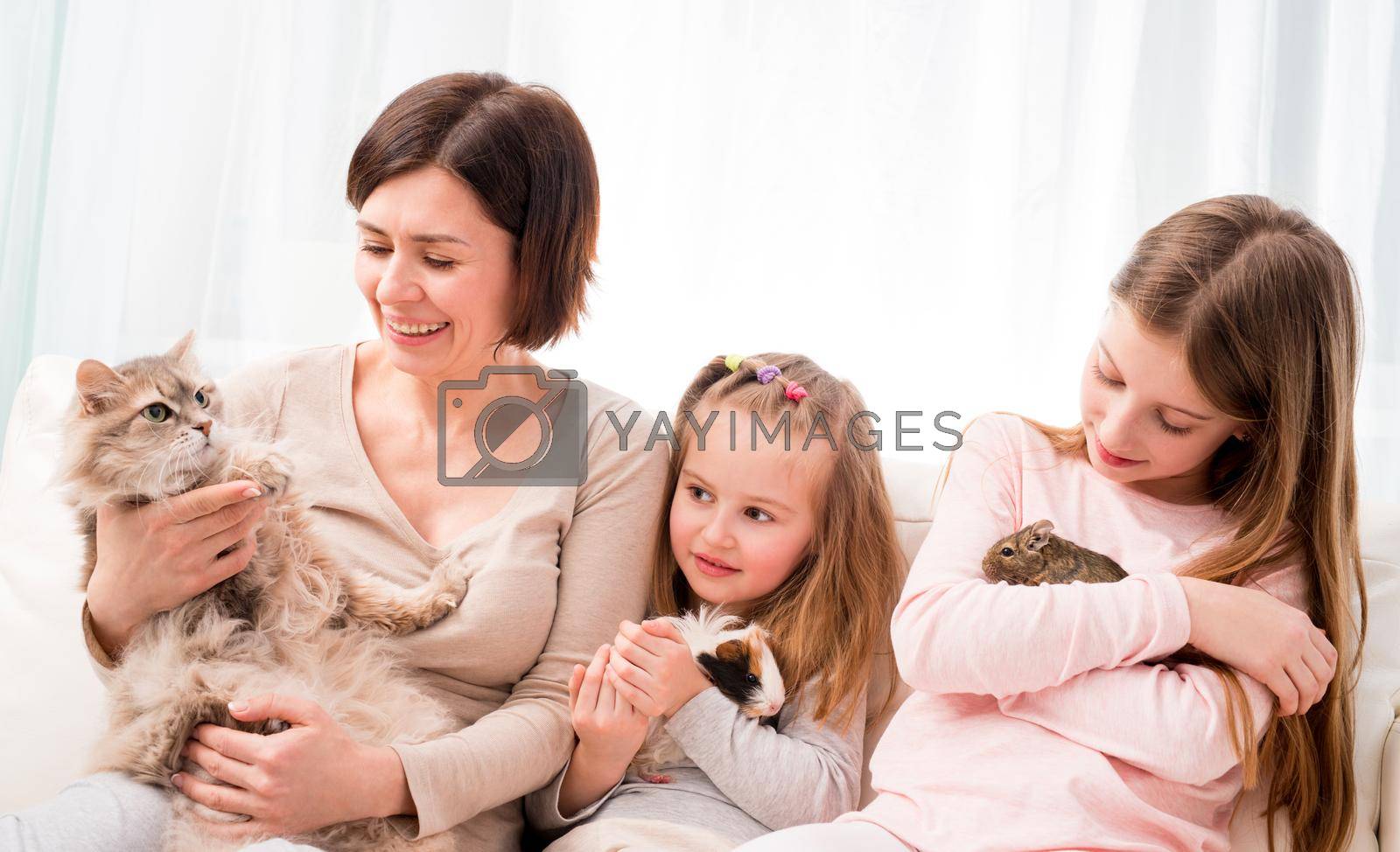 Mother and two little daughters holding their favorite pets on hands. Children playing with guinea pig, cat and degu squirrel while sitting on the soft sofa at home.