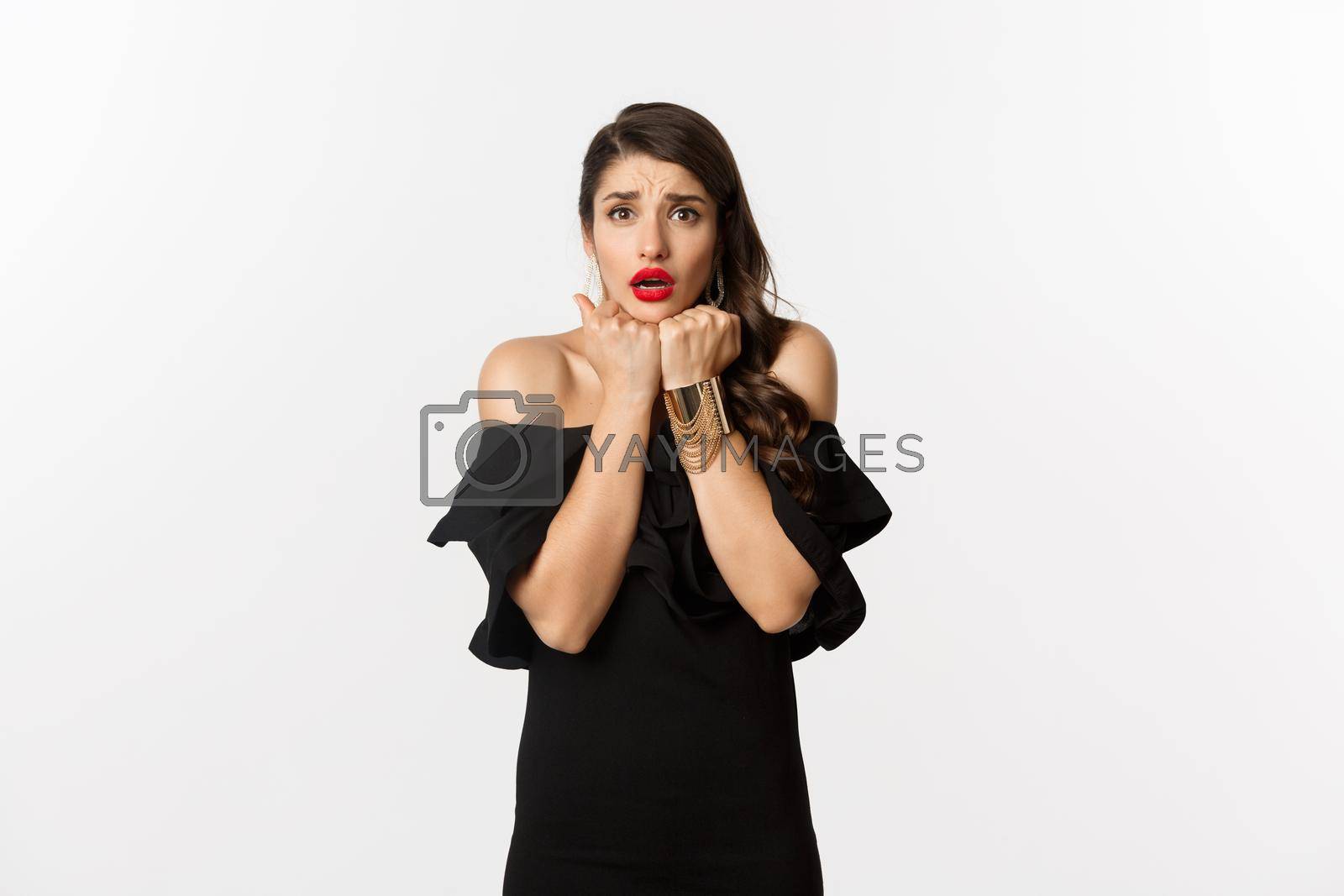 Royalty free image of Fashion and beauty concept. Scared timid woman grimacing, looking concerned and worried at camera, standing over white background in black dress by Benzoix