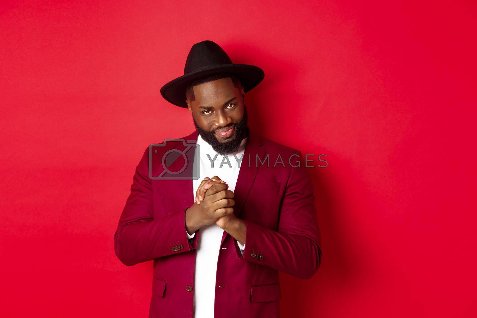 Royalty free image of Cunning black man having an idea, smiling devious and rubbing hands, standing against red background and scheming by Benzoix