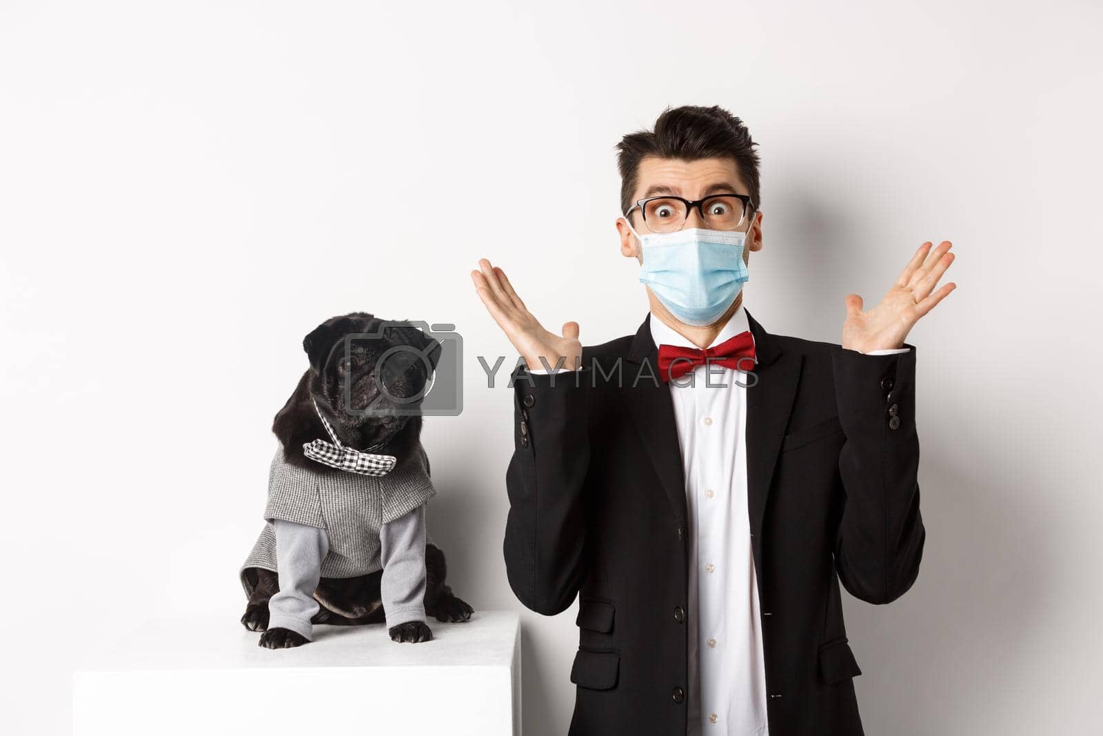Coronavirus, pets and celebration concept. Amazed young man in face mask and suit staring at camera surprised, cute black dog sitting near owner in party outfit, white background.