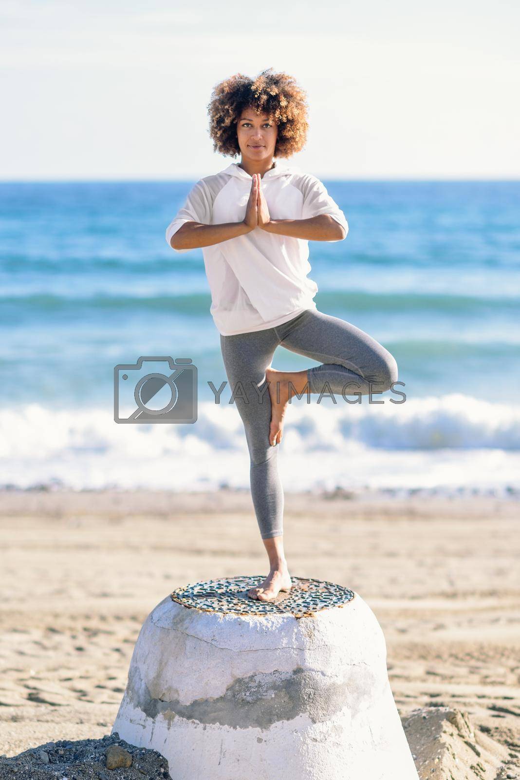 Young black woman doing yoga in the beach. Female wearing white sport clothes in tree asana with defocused sea background. Afro hairsytle.