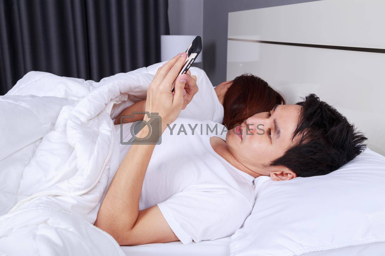 Royalty free image of man using his mobile phone in bed while his wife is sleeping next to him by geargodz