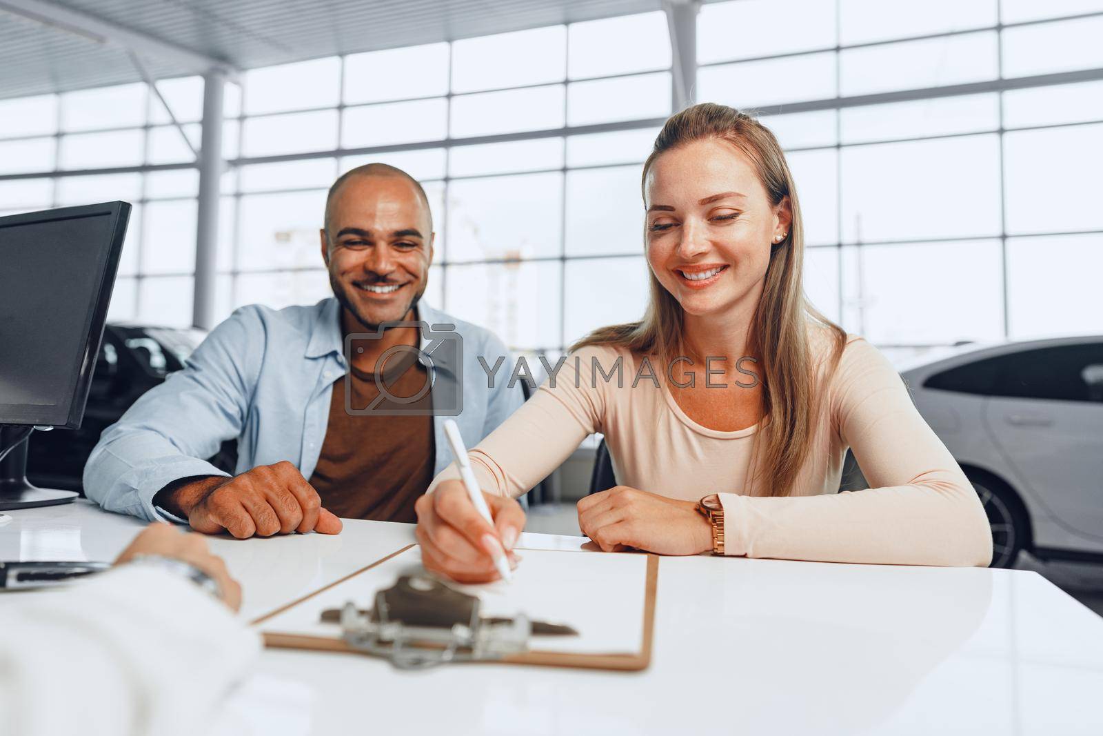 Royalty free image of Beautiful young couple signs documents at car dealership showroom by Fabrikasimf