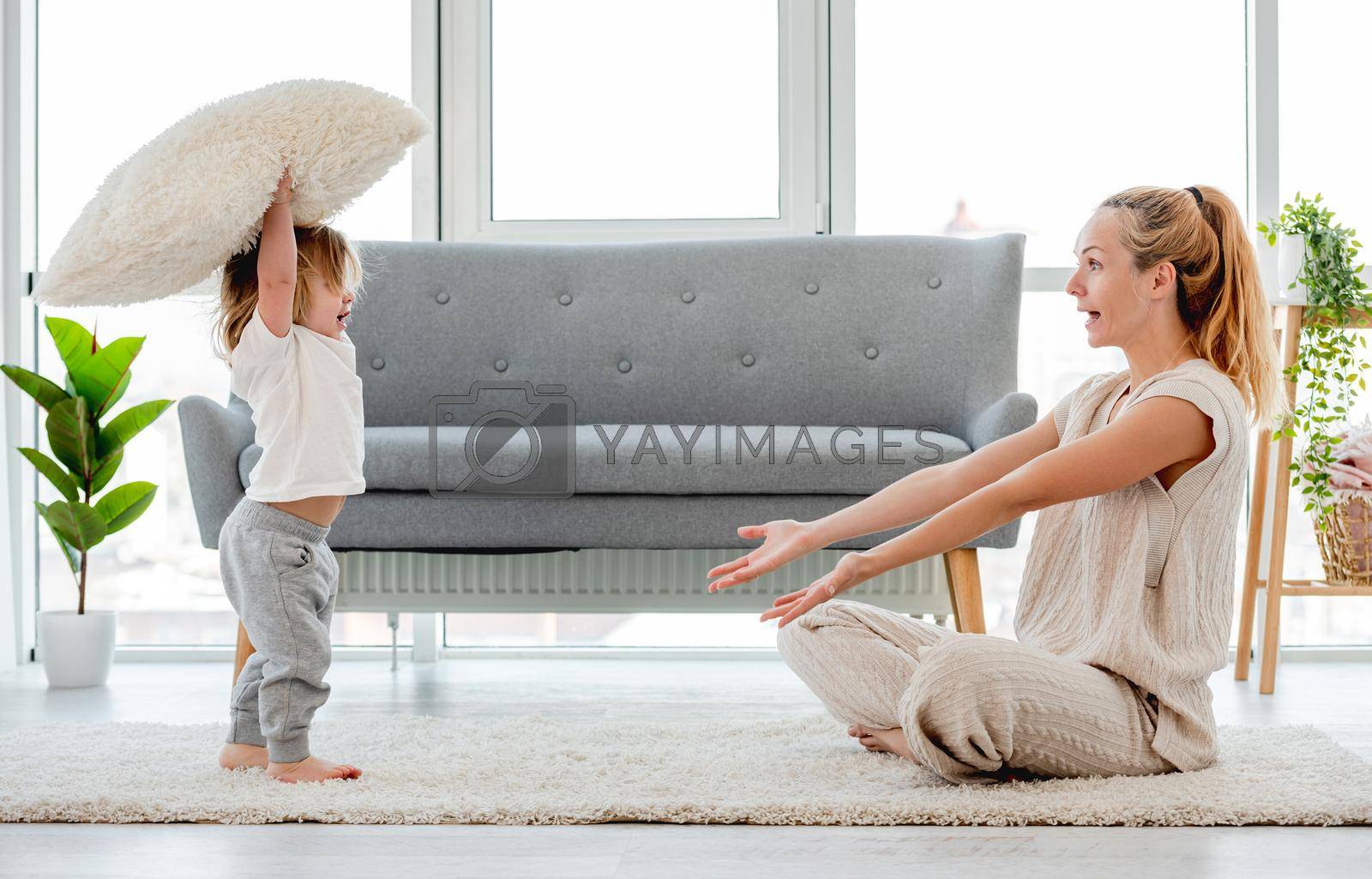 Beautiful blond young mother sitting on the floor and playing with her baby child son with pillow. Happy family portrait
