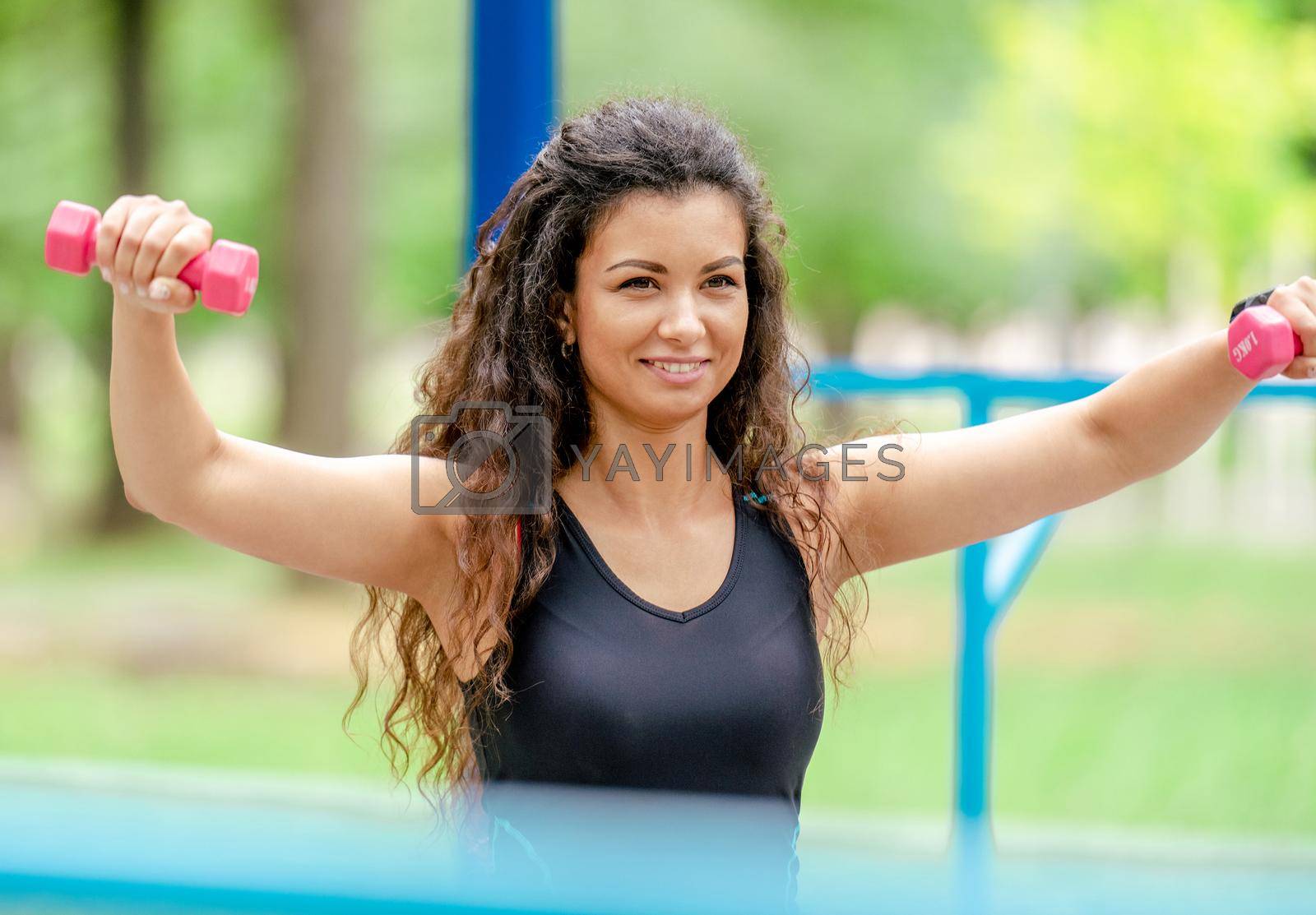 Pretty girl with dumbbells doing arm workout outdoors. Young woman exercising with sport fitness equipment at stadium