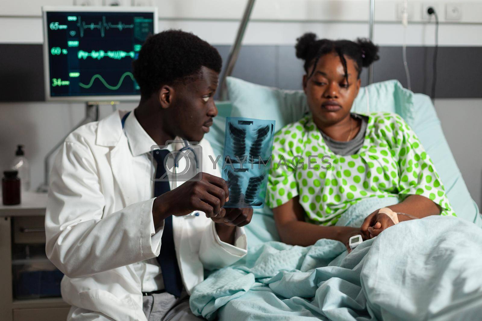 African american practitioner doctor holding lungs radiography explaining healthcare treatment to young woman during clinical appointment in hospital ward. Patient recovering after surgery