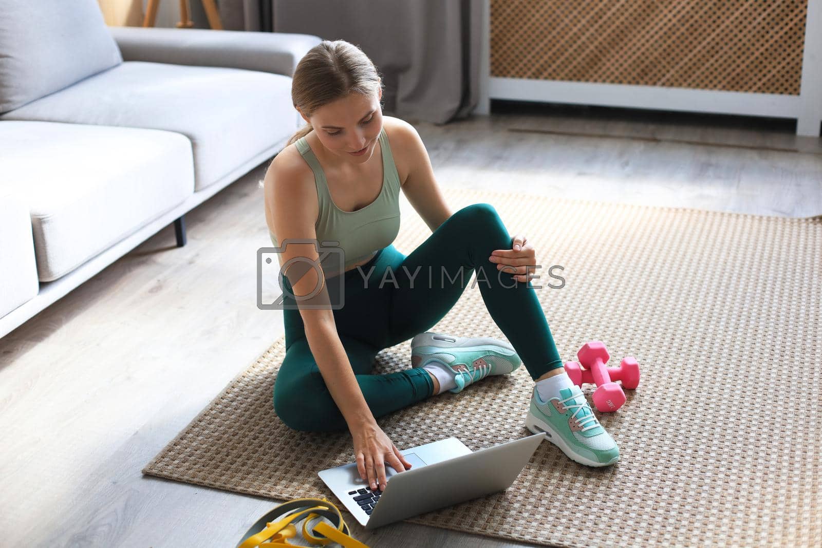 Royalty free image of Beautiful slim sporty woman in sportswear is sitting on the floor with dumbbells and is using a laptop at home in the living room. Healthy lifestyle. Stay at home activities. by tsyhun