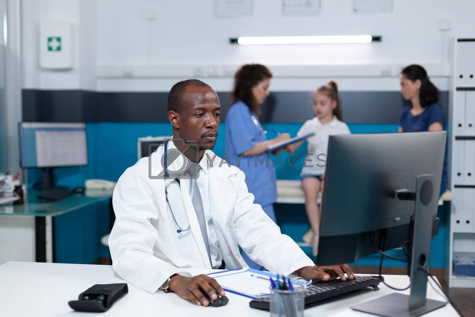 African american pediatrician doctor working at healthcare treatment in hospital office typing medical expertise on computer during clinical examination. In background nurse examining patient