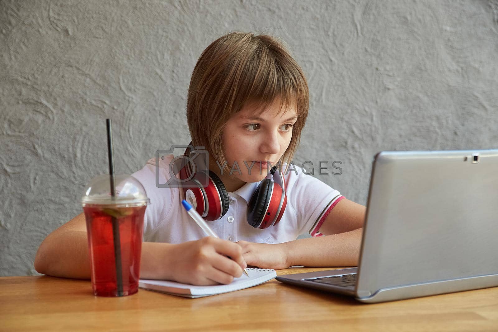 schoolgirl with headphones sits at table, watching at laptop screen, writing notes at copybook. little caucasian girl studying with computer, doing homework. distant learning, online education, lesson