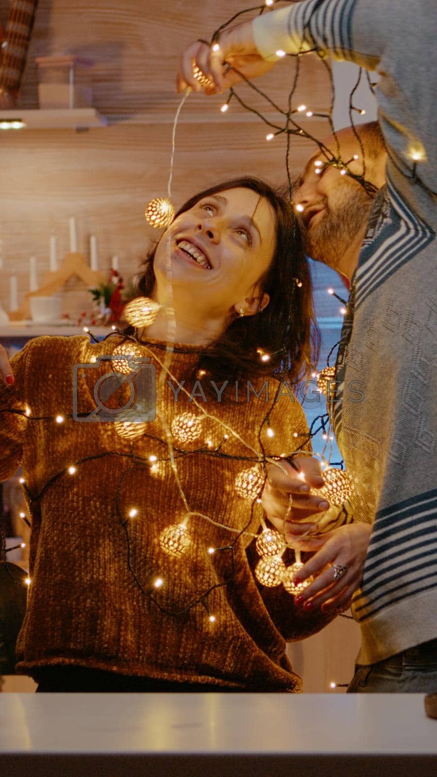Cheerful people getting tangled in festive lights while decorating home for christmas eve celebration. Man and woman smiling, trying to untangle garland of twinkle lights bulbs.