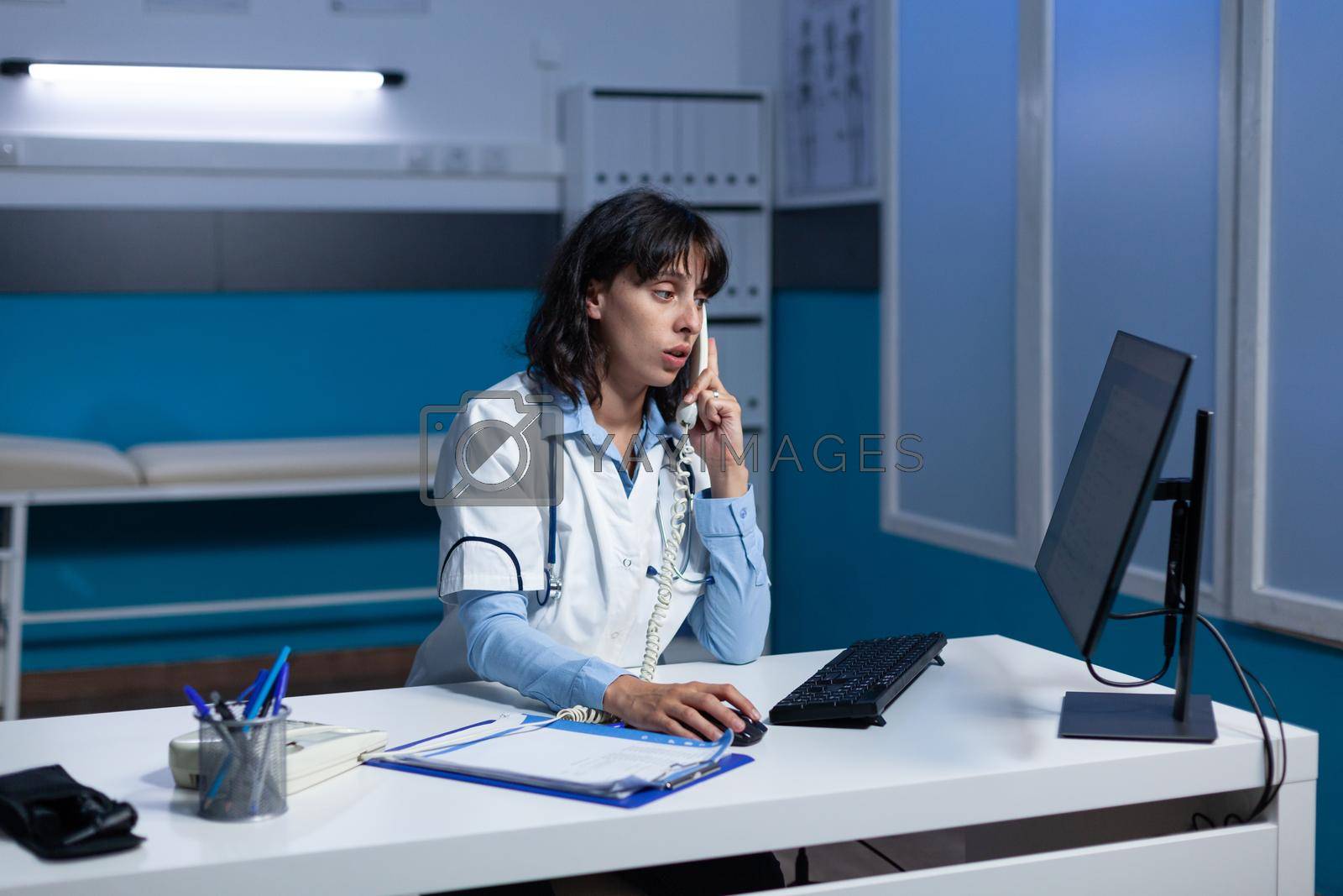 Doctor talking on landline phone for healthcare checkup at medical office. Woman working as medic using telephone for remote conversation about appointment with patient, working late.