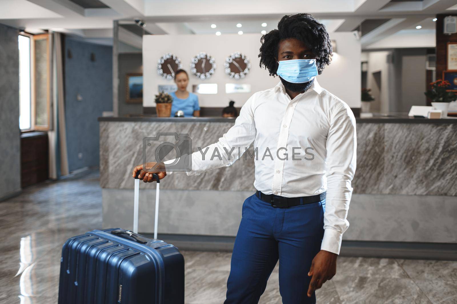 Royalty free image of African man hotel guest with suitcase wearing protective mask to protect from coronavirus by Fabrikasimf