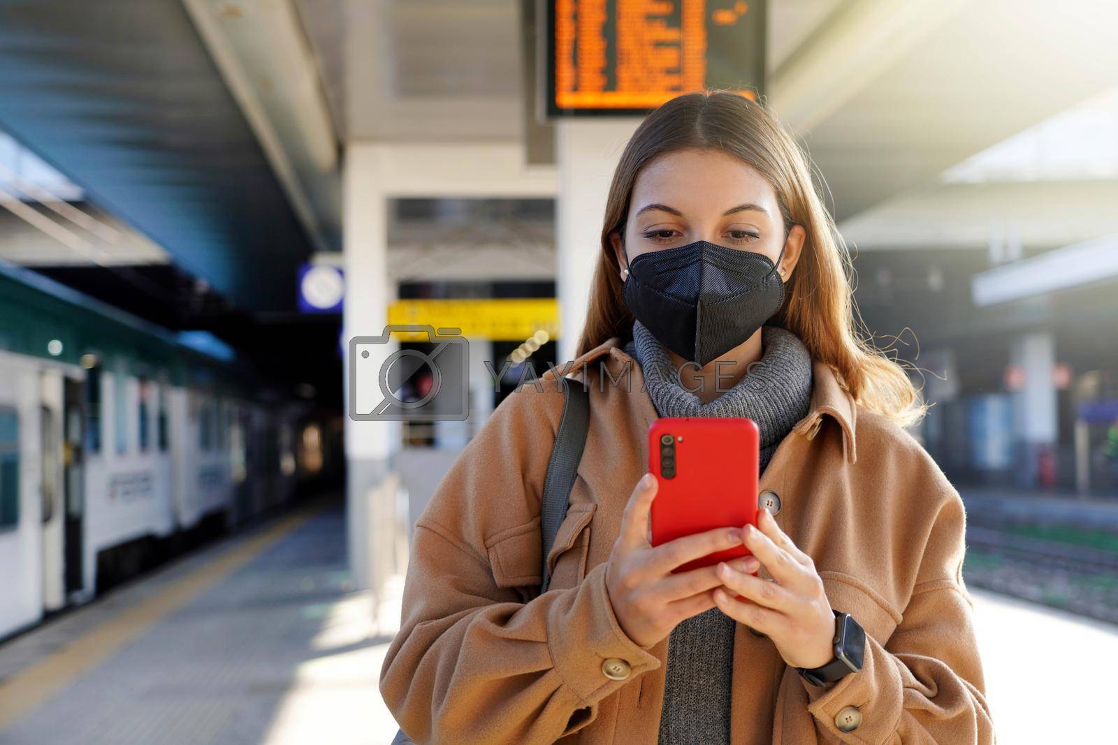 Stylish urban woman wearing black face mask KN95 FFP2 texting on smartphone in train station