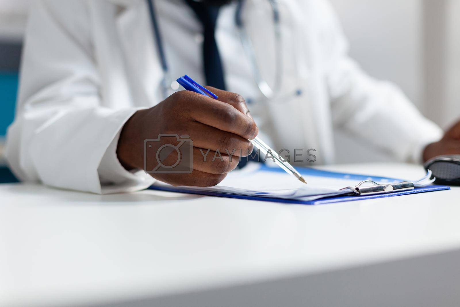 Closeup of african american doctor hand writing healthcare treatment on paperwork after analyzing medicine prescription. Therapist analyzing sickness symptoms expertise in hospital office
