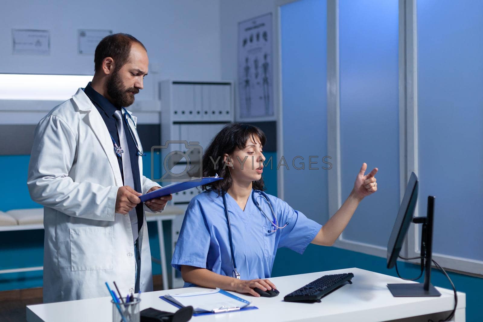 Medical team of specialists looking at computer for healthcare information and examination. Doctor holding checkup files while nurse working with monitor on desk. Man and woman at job