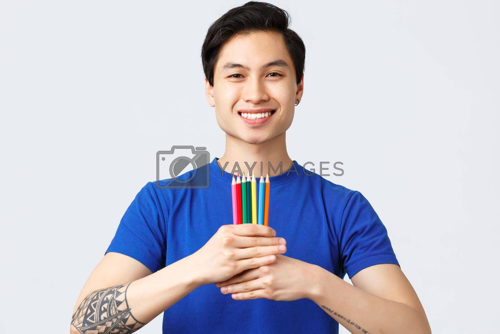 Self-quarantine, social distancine, leisure and lifestyle concept. Young handsome asian man with tattoo show colored pencils, found new hobby feeling happy, smiling at camera.