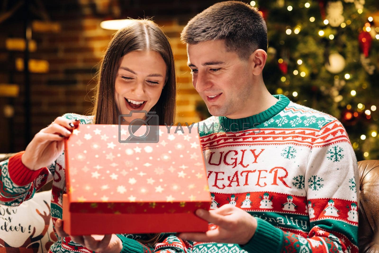 Royalty free image of Portrait of cheerful young woman opening xmas gift box with excited surprised face feeling happy on New Year's Eve sitting at home near glowing christmas tree. Xmas concept. Holiday miracle by uflypro