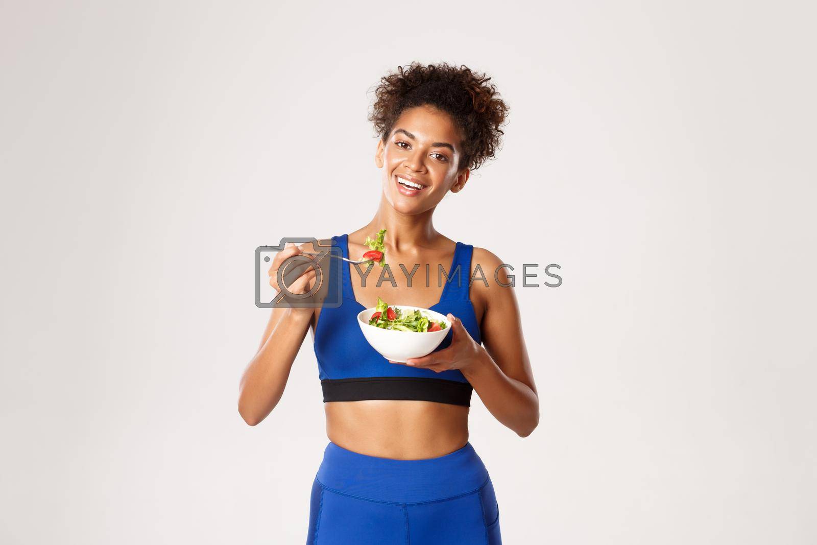 Healthy lifestyle and sport concept. Attractive young fitness woman in blue sport outfit, eating salad and smiling, standing over white background.