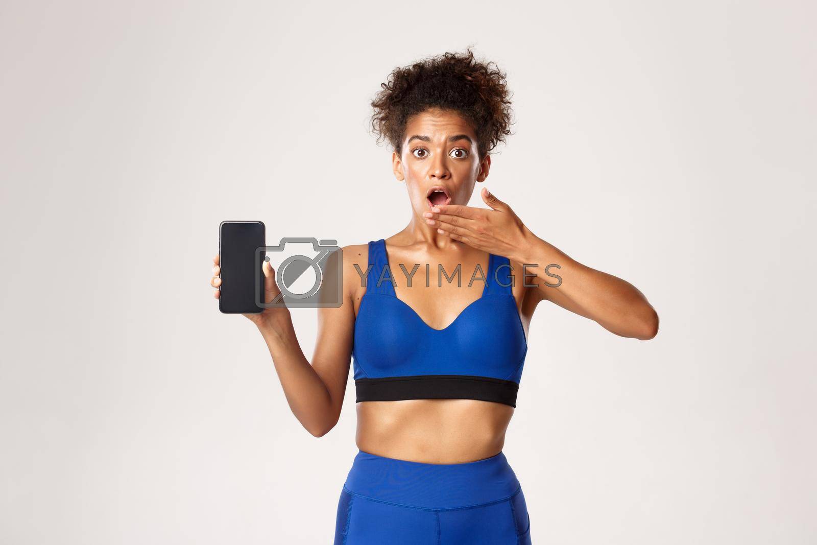 Sport and technology concept. Surprised african-american sportswoman in fitness clothing, gasping amazed, showing mobile phone screen, white background.